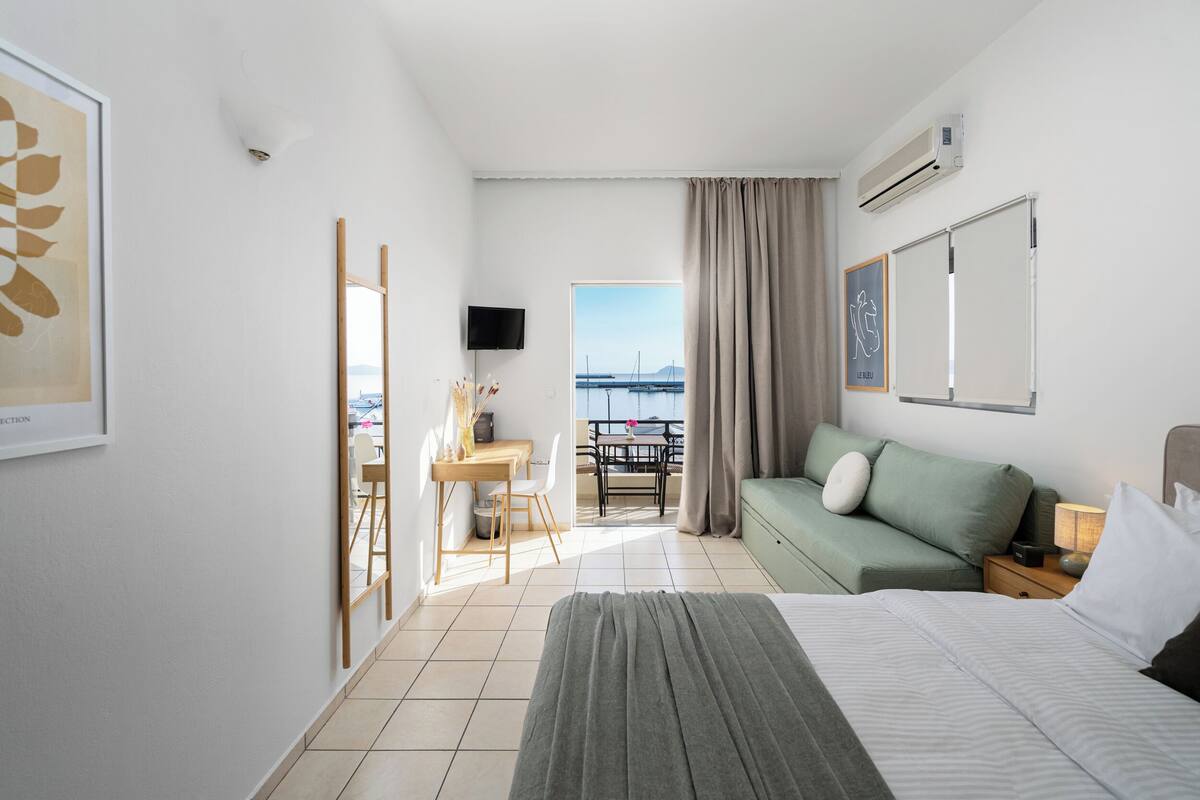 Habitación de hotel con cama, sofá, escritorio con sillas, televisor y balcón con vista al mar.