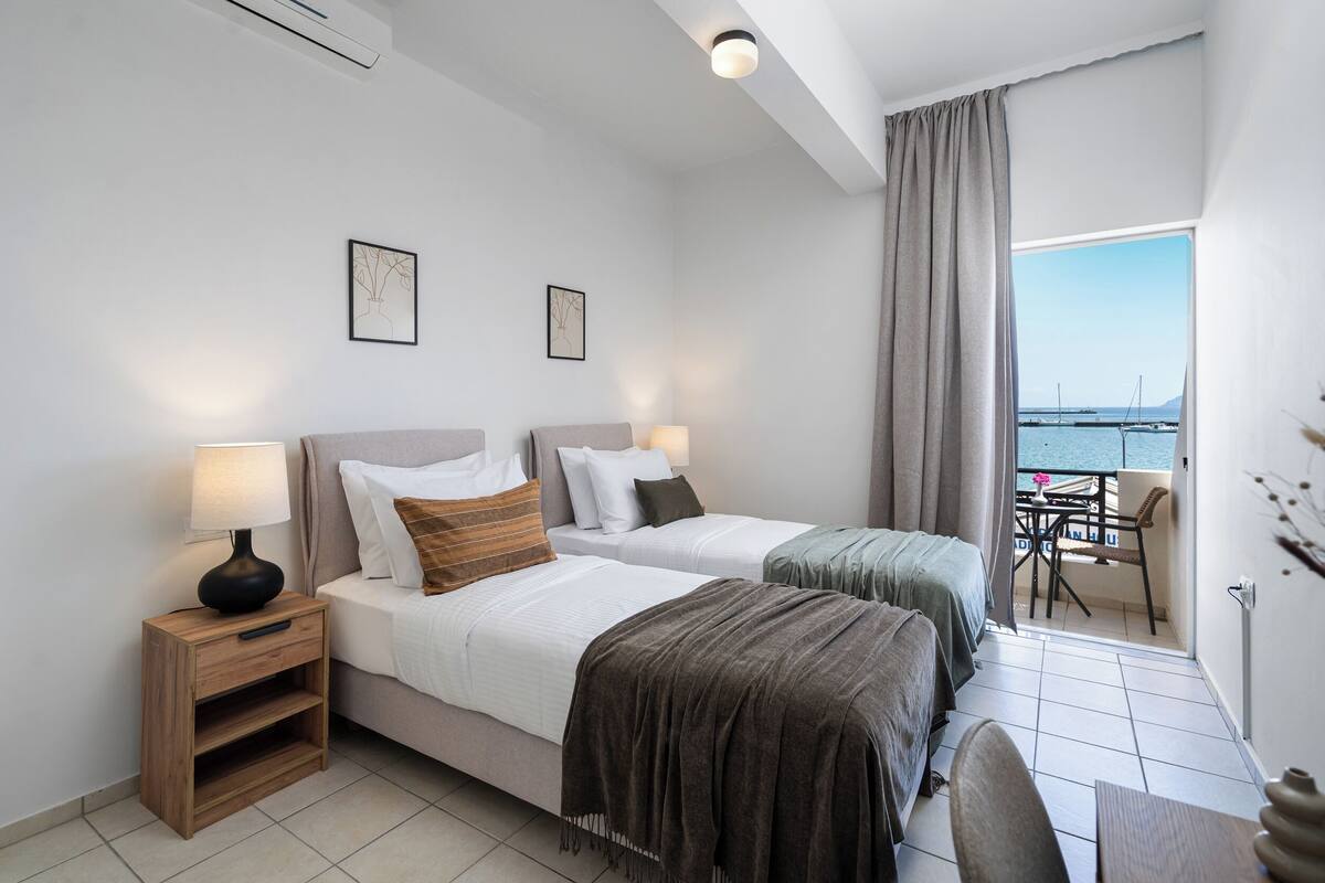 Habitación de hotel con dos camas, una mesita de noche, una silla, una mesa y vista al mar.