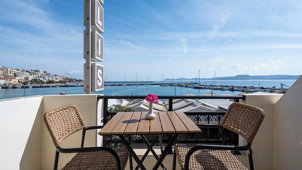 A balcony with wicker chairs, a wooden table, and a vase with pink flowers. - Hotel Flisvos (Sitia)