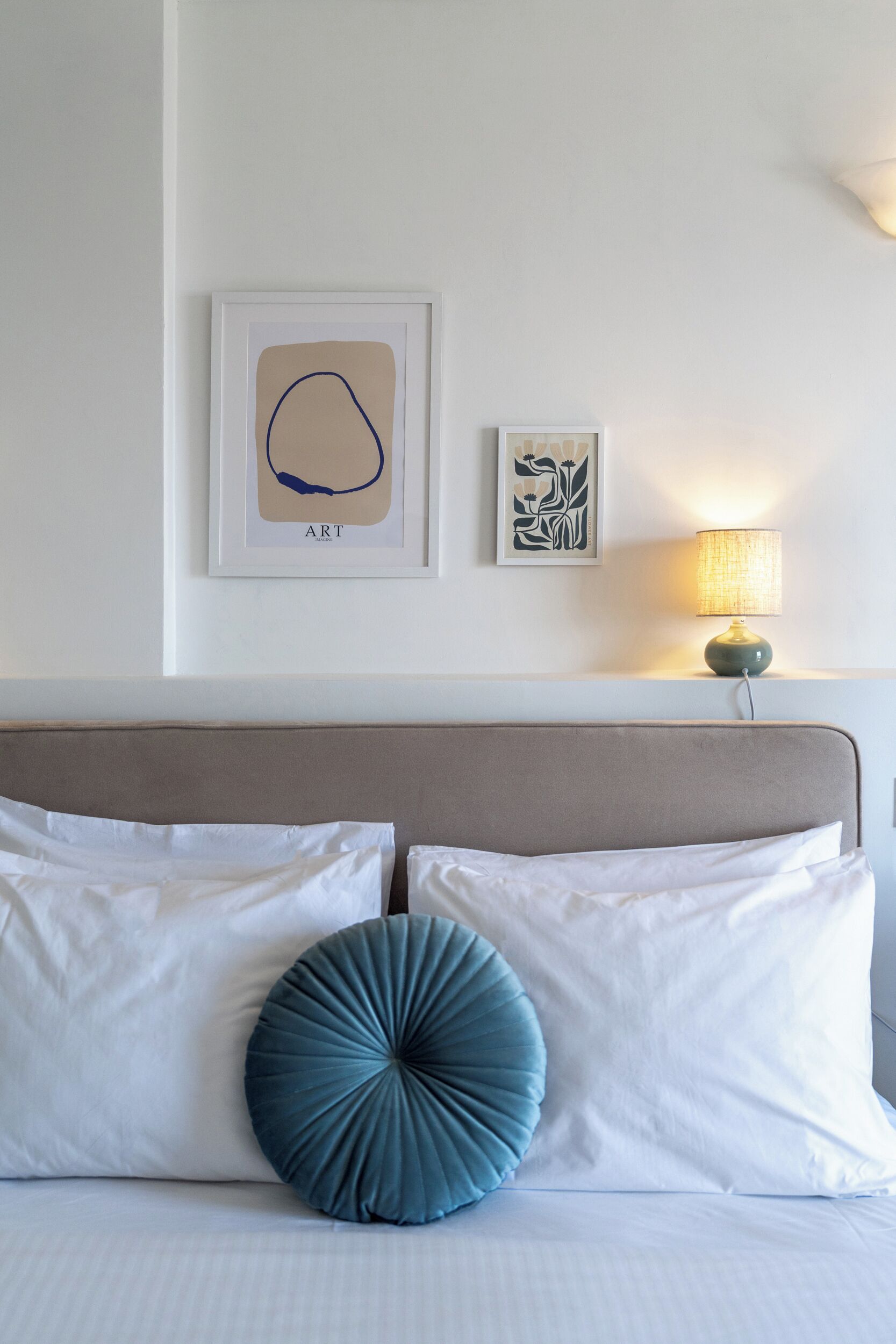 Una cama bien arreglada con sábanas blancas y una única almohada azul decorativa, una lámpara de noche y dos cuadros en la pared.