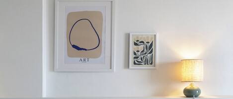 Una cama bien arreglada con sábanas blancas y una única almohada azul decorativa, una lámpara de noche y dos cuadros en la pared.