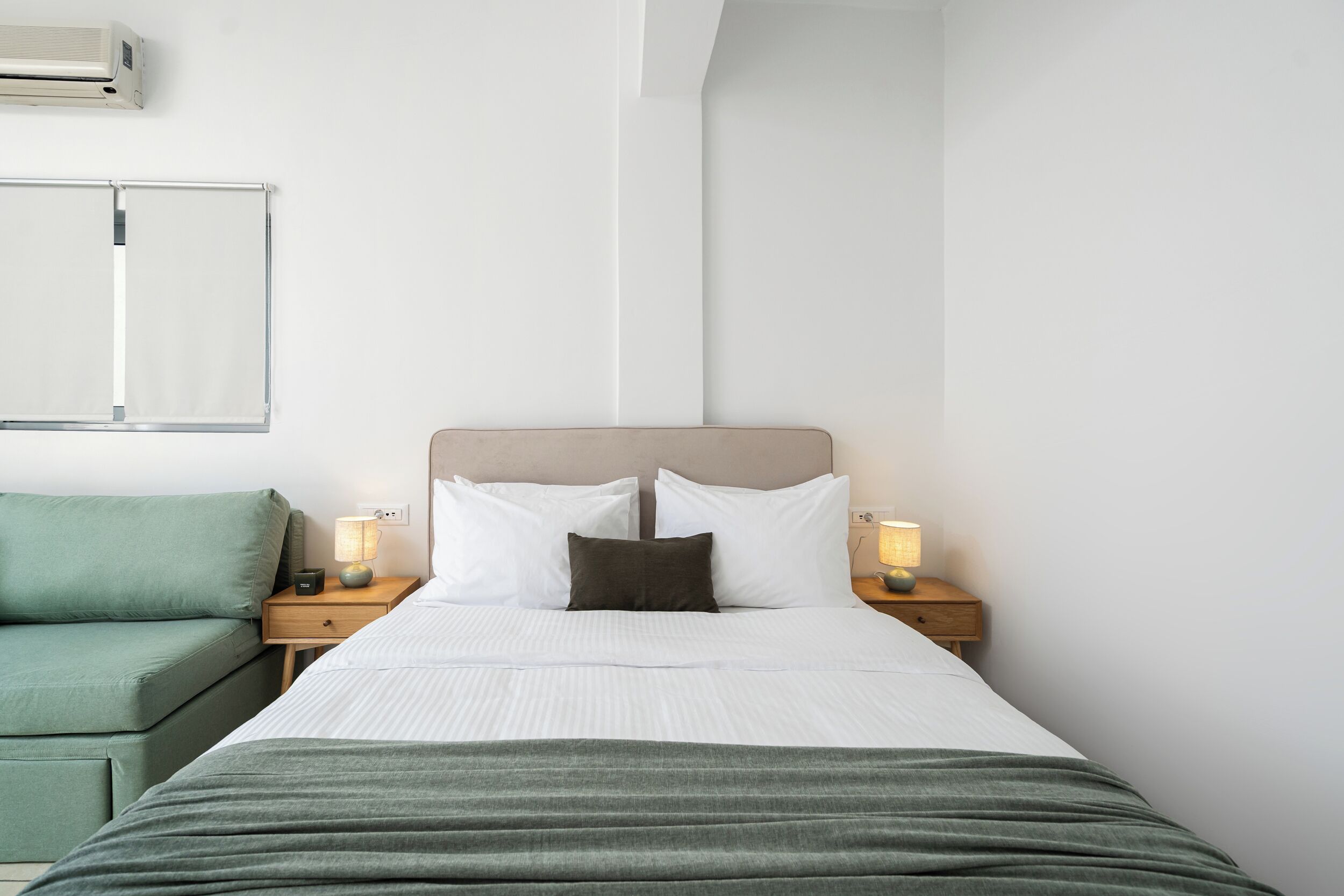 A hotel room with a bed, a green sofa, two bedside tables with lamps, and a wall-mounted air conditioner.