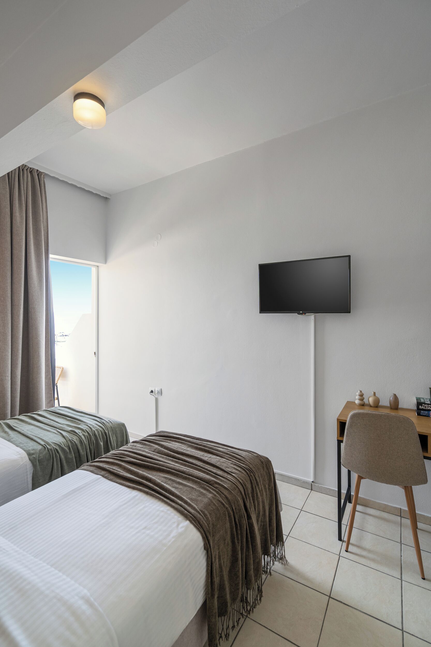 Habitación de hotel con dos camas, un televisor montado en la pared, un escritorio con silla y una ventana con cortinas.