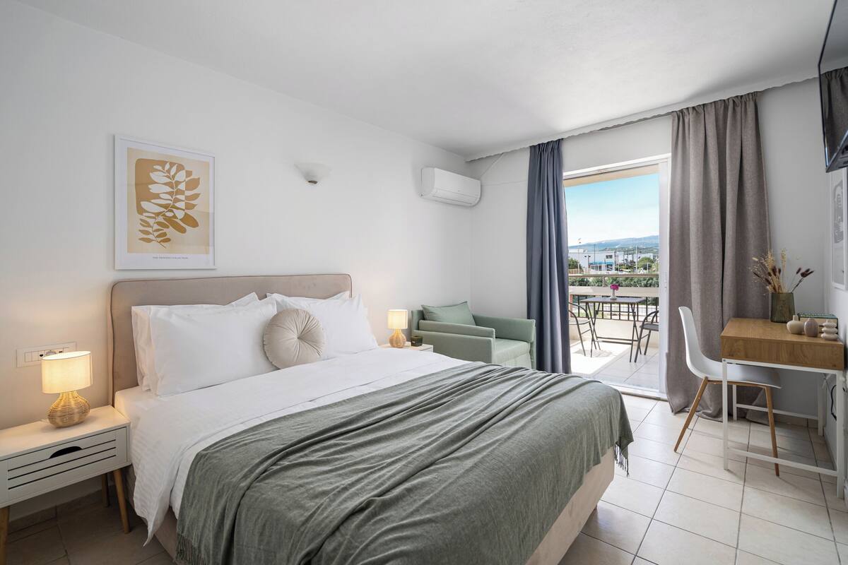 Habitación de hotel con cama, mesita de noche, lámpara y vistas al exterior.