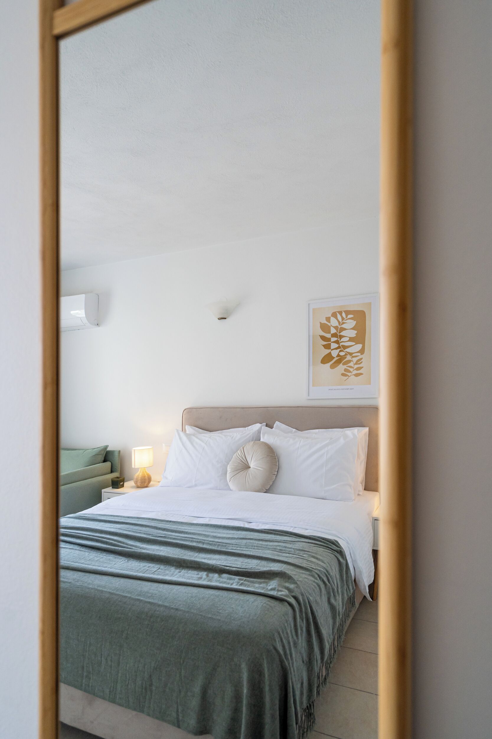 A bedroom with a bed, a nightstand, a lamp, a wall-mounted air conditioner, and a framed artwork.