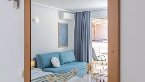 A room with a bed, a blue sofa, a small table with a vase, and a mirror on the wall. - Hotel Flisvos (Sitia)