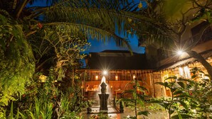 Un jardín exterior bien iluminado con una fuente central, rodeado de exuberante vegetación tropical y un edificio de arquitectura tradicional.