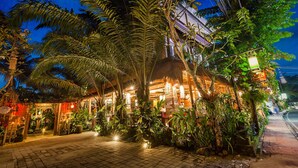 Un espacio exterior bien iluminado con un restaurante, rodeado de abundante vegetación y palmeras.