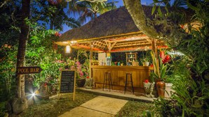 Un bar junto a la piscina con techo de paja, rodeado de exuberante vegetación tropical y un cartel con el nombre del bar.