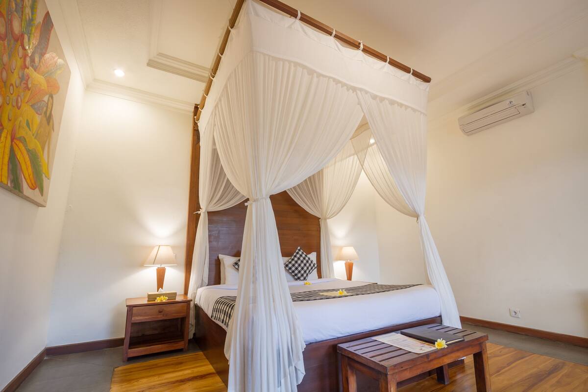 A four-poster canopy bed with white linens, two bedside tables with lamps, and a wooden floor.