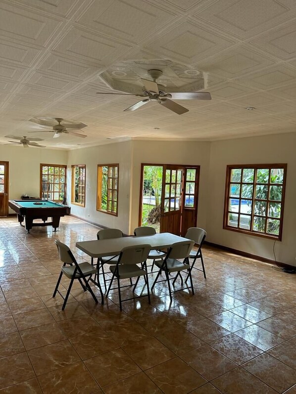 A spacious indoor area with a pool table, a dining table with chairs, and large windows with views of greenery. - Brillasol Airport Hotel (San Antonio)