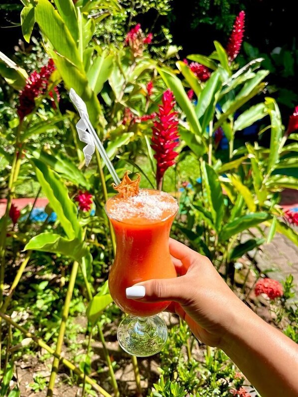 A hand holding a glass with a red drink and a decorative garnish, set against a backdrop of lush greenery and red flowers. - Brillasol Airport Hotel (San Antonio)