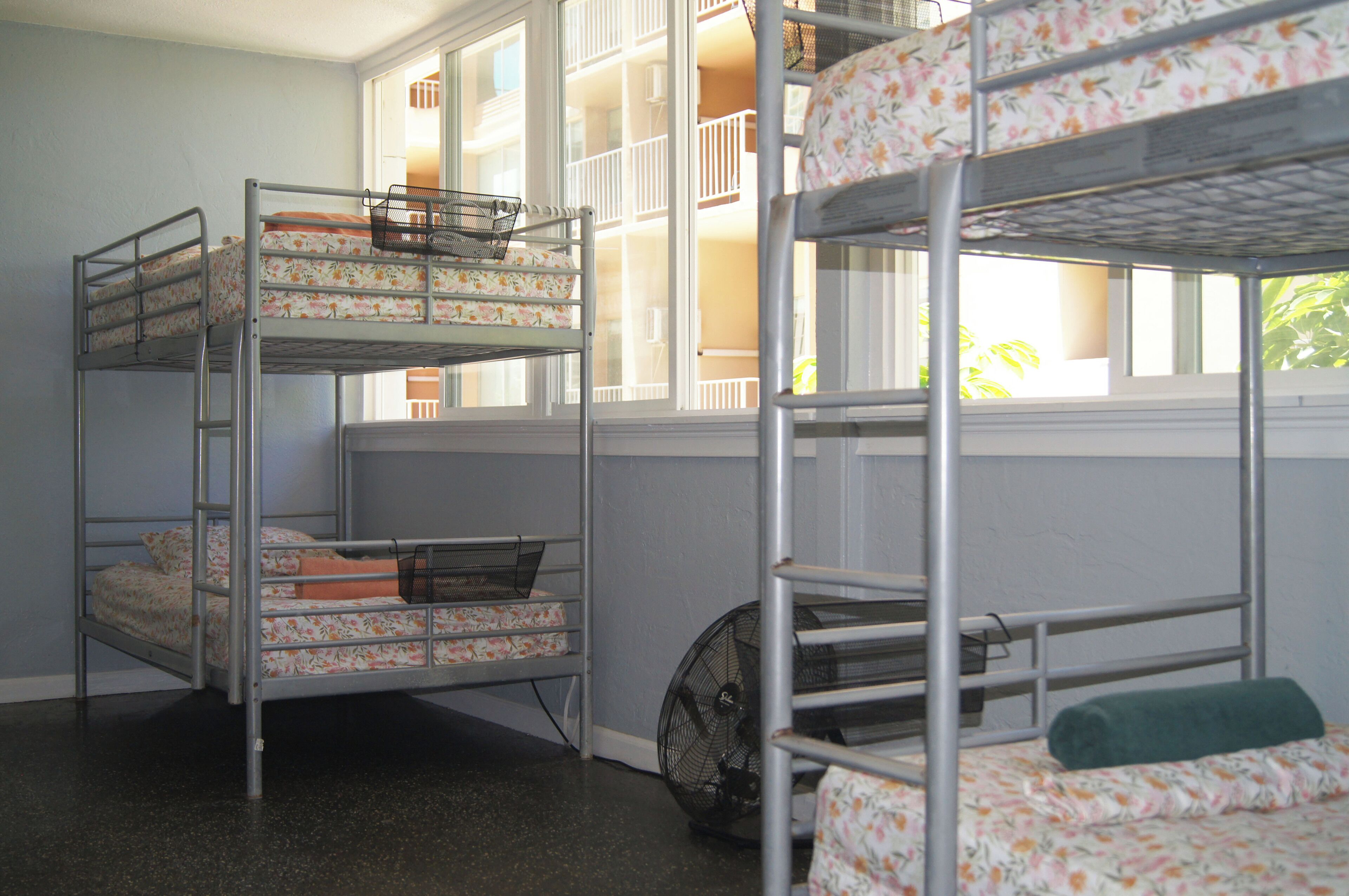 Dormitorio con literas, varios catres, un ventilador y una ventana que da a balcones.