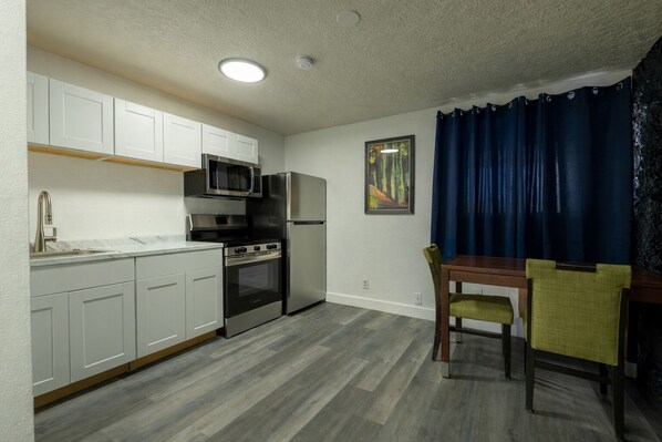 A compact kitchen with white cabinets, a microwave, a stove, and a refrigerator. There's a dining table with chairs and a blue curtain.