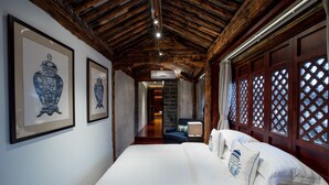 Une chambre d’hôtel avec un plafond en bois, un grand lit recouvert de draps blancs, un vase bleu et blanc accroché au mur, et une vue à travers une fenêtre à croisillons.