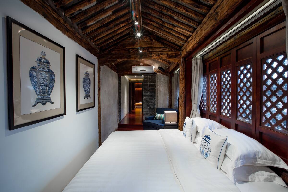 Ein Hotelzimmer mit Holzboden, einem großen Bett mit weißen Leinen, einer blauen und weißen Vase an der Wand und Blick durch ein Gitterfenster.