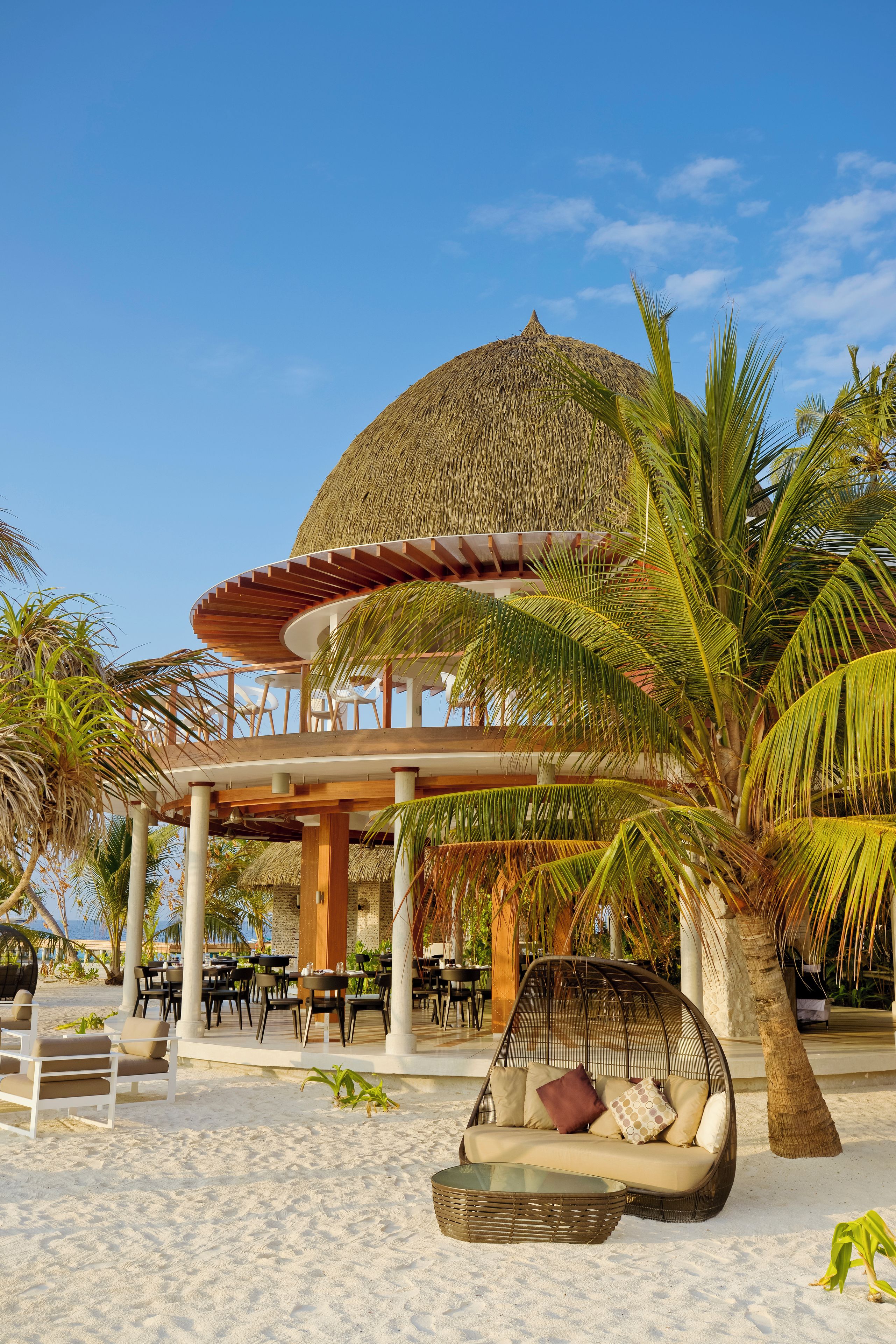 Eine Strandlounge mit einem Reetdach, Sitzgelegenheiten im Freien und Palmen.
