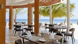 En restaurang vid stranden med bord och stolar, palmer och utsikt över havet.