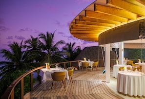 Un área de comedor al aire libre con mesas y sillas, rodeada de palmeras y una terraza de madera.