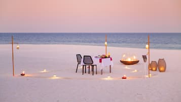 A beach dinner setup with a table, chairs, candles, and lanterns.