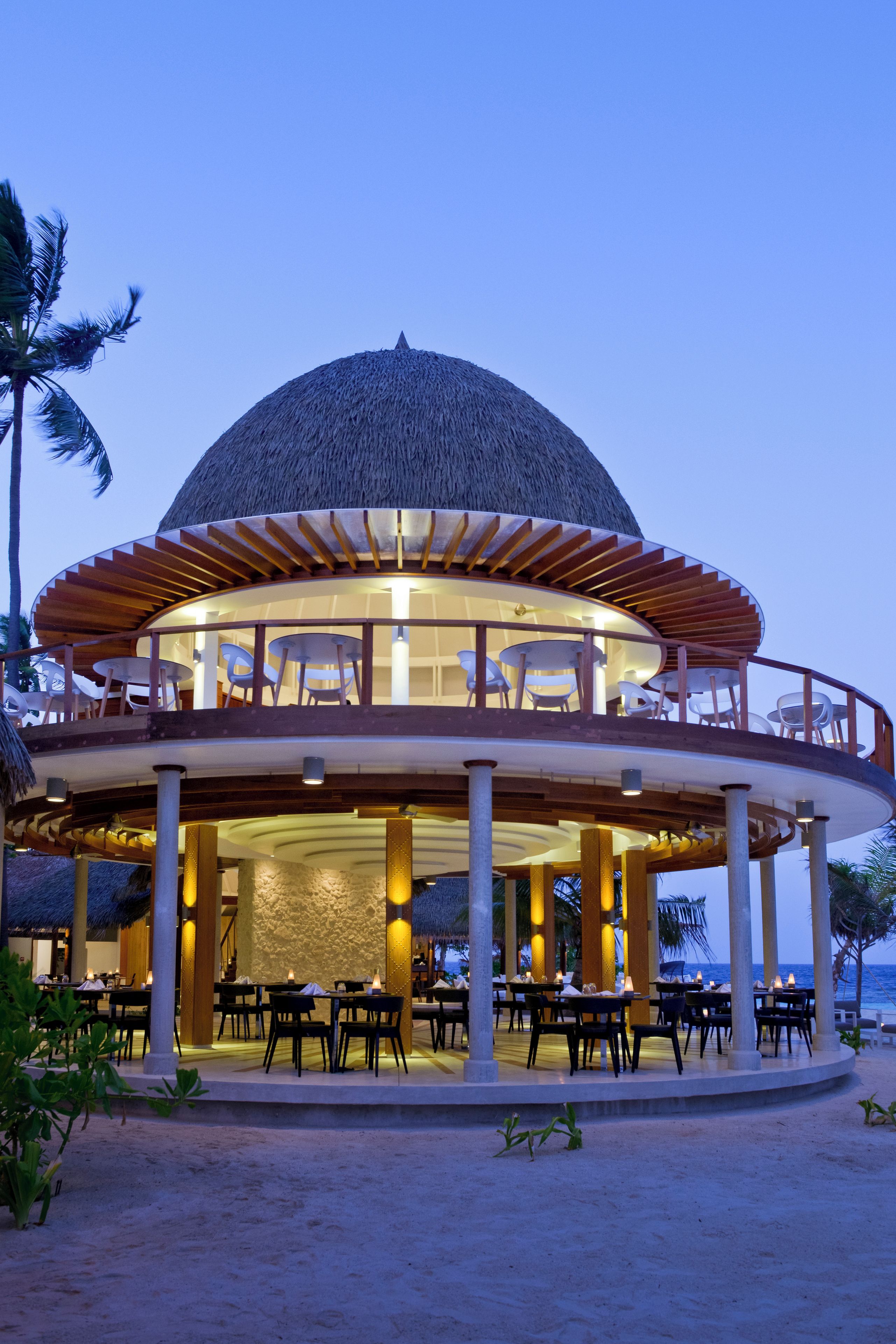 Un restaurante en la playa con techo de paja, mesas al aire libre y vista al océano.