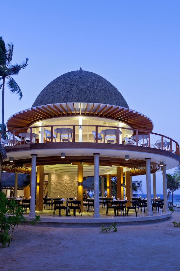 Un restaurante en la playa con techo de paja, mesas al aire libre y vista al océano.