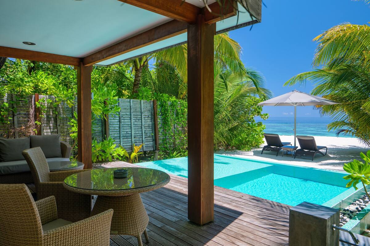 Una terraza de madera con una mesa de vidrio y sillas de mimbre, con vista a una piscina y una playa.
