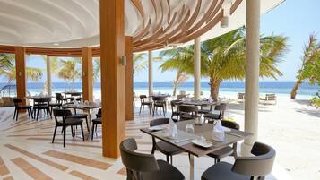 A beachside restaurant with tables and chairs, palm trees, and a view of the ocean.