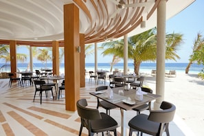 A beachside restaurant with tables and chairs, palm trees, and a view of the ocean. - Kandolhu Maldives (NORTH ARI ATOLL)