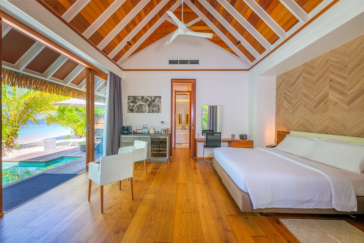 Une chambre avec un plafond en bois, un lit, un bureau et une vue sur une piscine et la verdure.