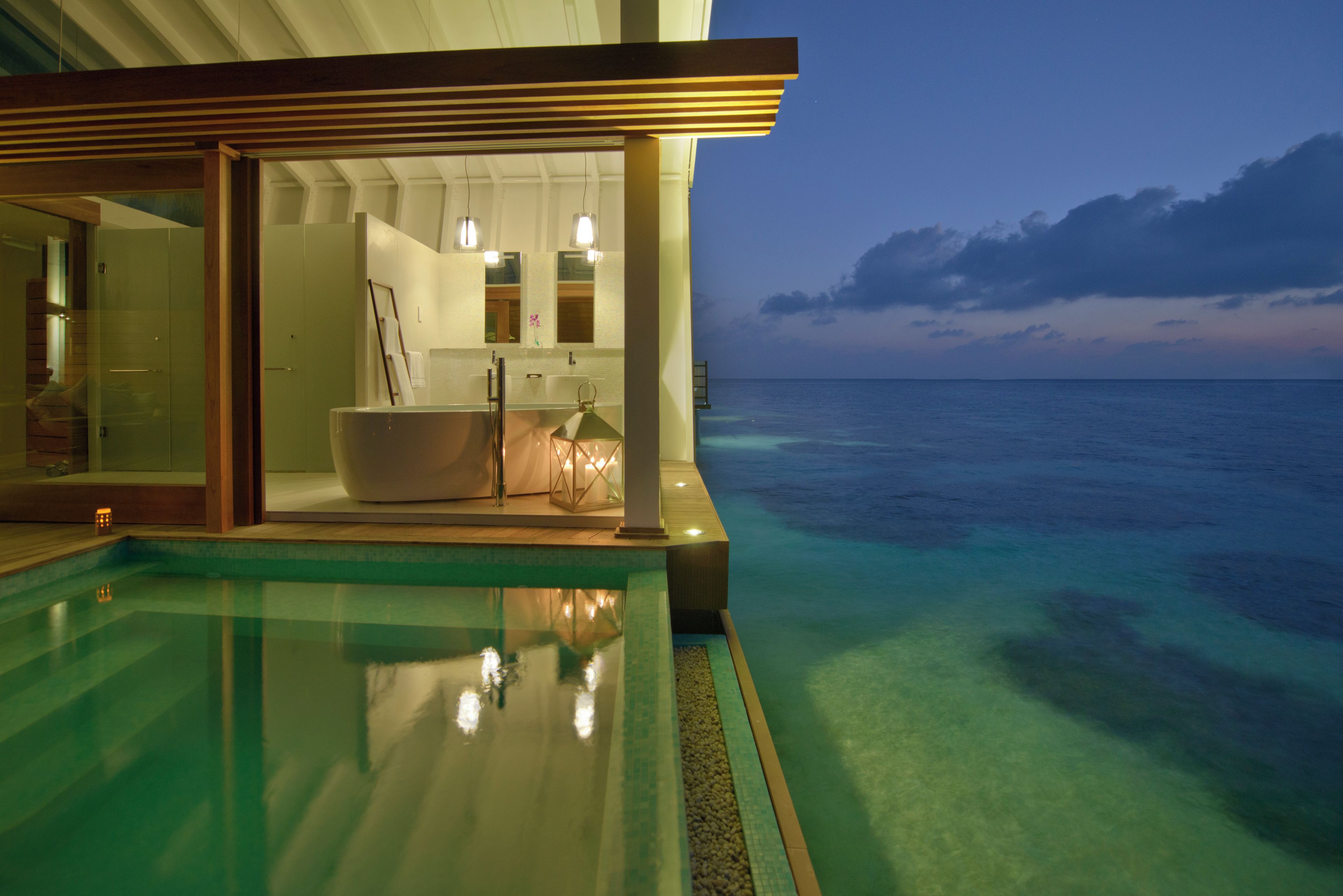Una piscina infinita moderna con agua marina cristalina, un baño privado con bañera y vistas al océano al atardecer.