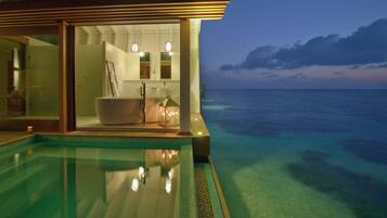 Una piscina infinita moderna con agua marina cristalina, un baño privado con bañera y vistas al océano al atardecer.