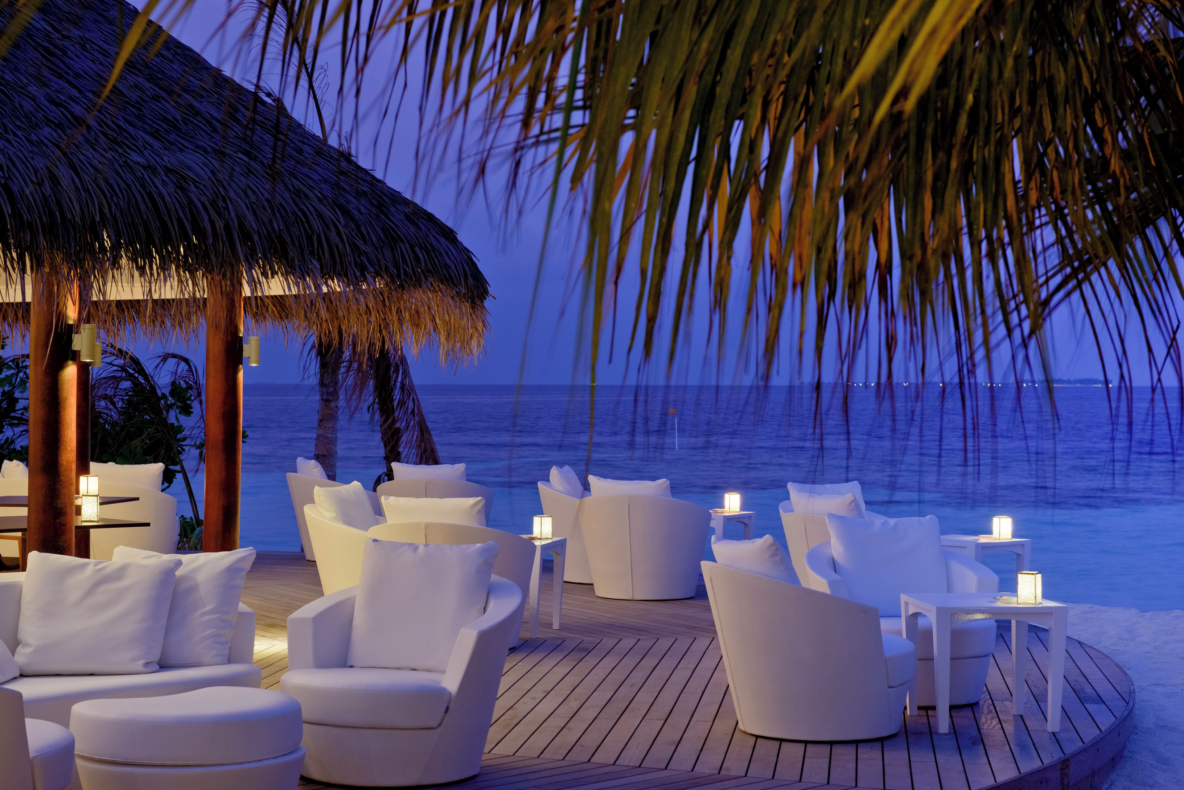 A deck with white sofas and tables, a thatched roof, and a view of the ocean.