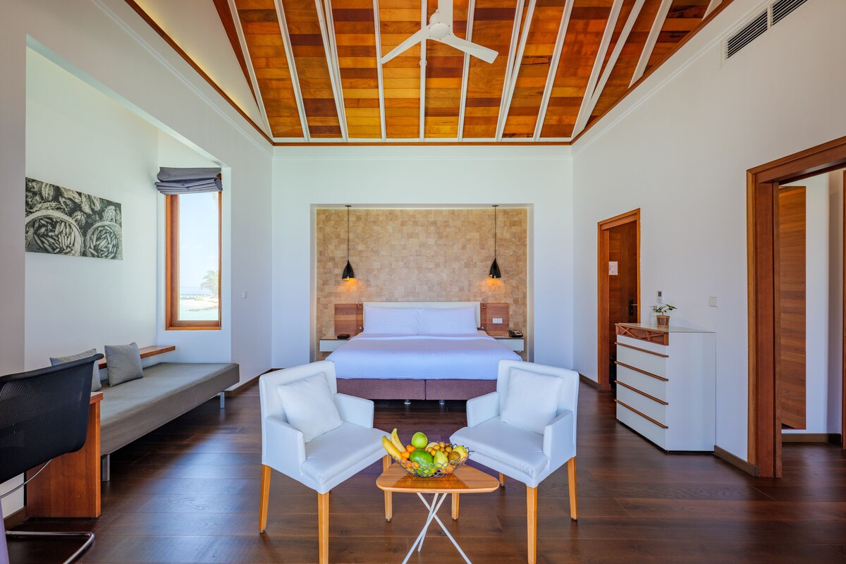 Une chambre d’hôtel moderne avec un grand lit, un bureau, deux chaises blanches, une petite table avec des fruits et un ventilateur de plafond.