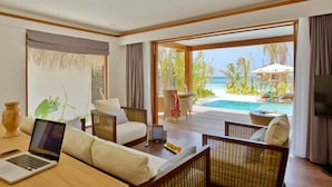 Ett hotellrum med en bärbar dator på ett bord, en soffa och utsikt över en pool och en strand.