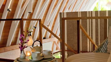 Un espacio circular y acogedor con techo de madera y una lámpara colgante. Hay una pequeña mesa con un hervidor, tazas y una vela. El ambiente ofrece una zona de estar confortable con cojines.