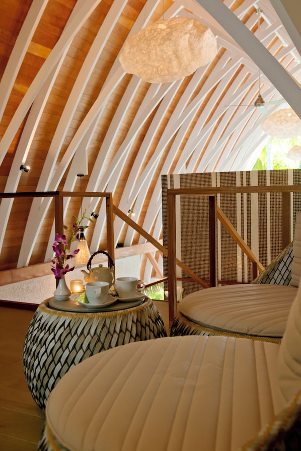 Un espacio circular y acogedor con techo de madera y una lámpara colgante. Hay una pequeña mesa con un hervidor, tazas y una vela. El ambiente ofrece una zona de estar confortable con cojines.