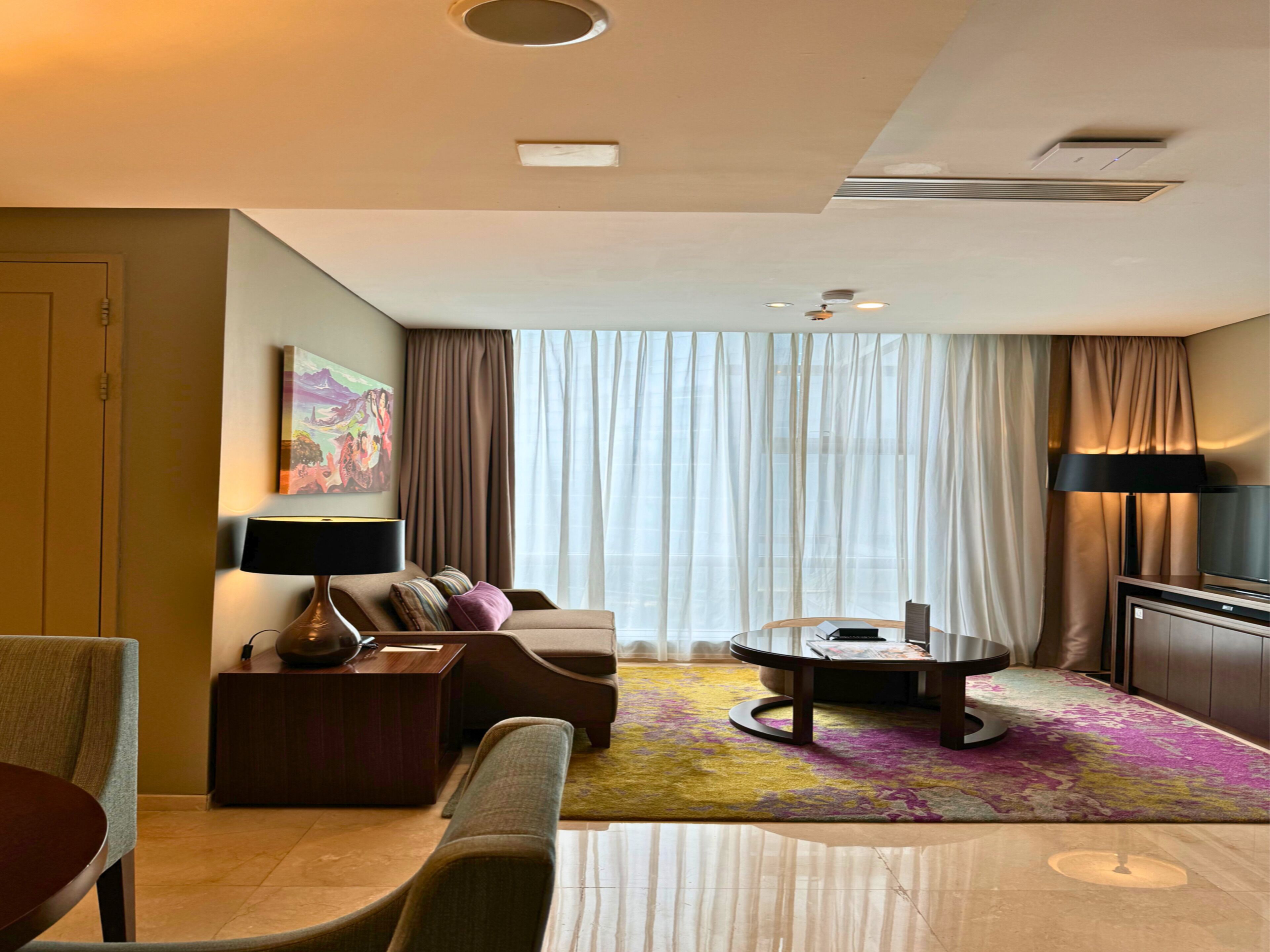 A modern hotel room with a sofa, a coffee table, a TV, and a large window with curtains.