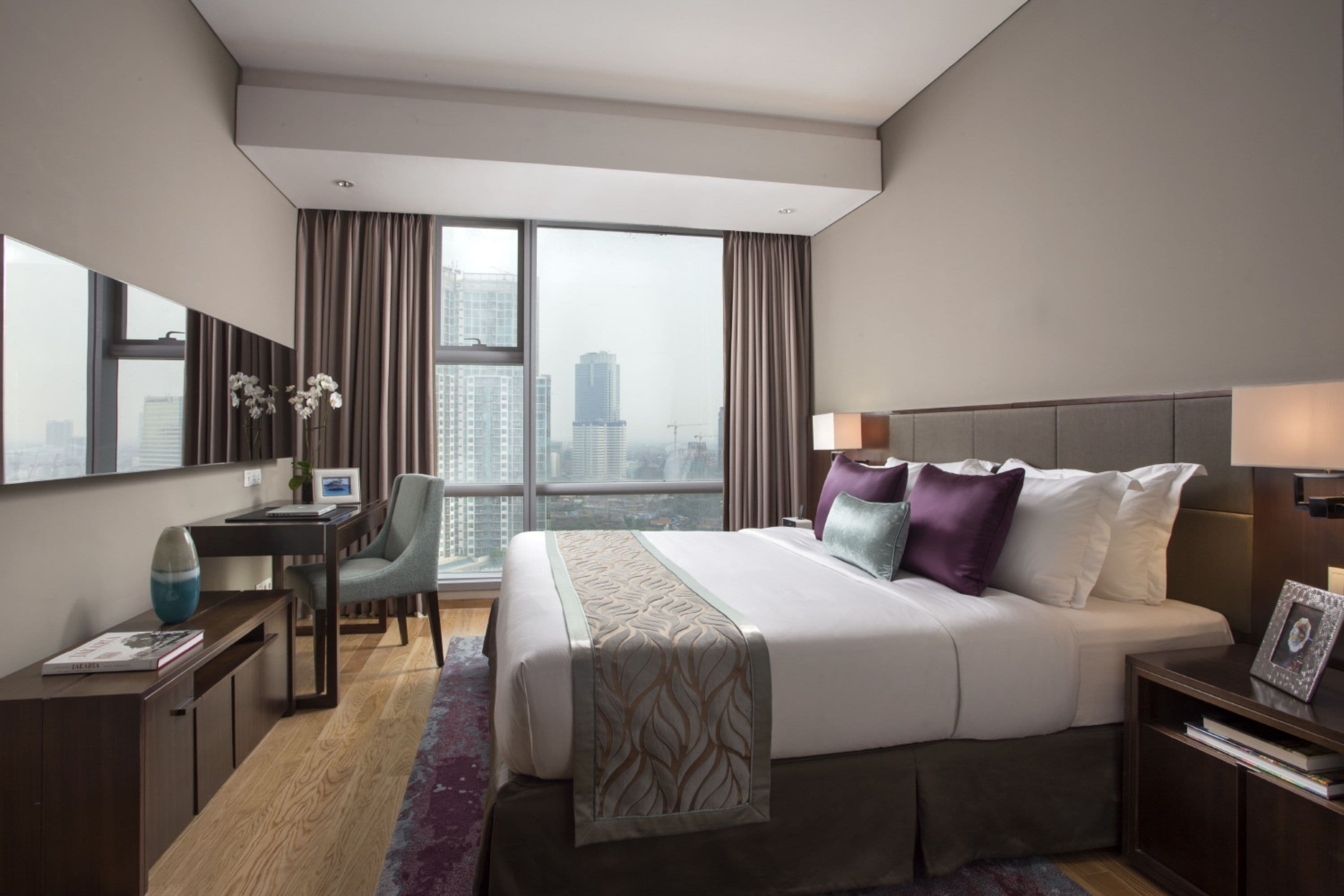 A modern hotel room with a large bed, a desk, a chair, and a view of the cityscape through large windows.