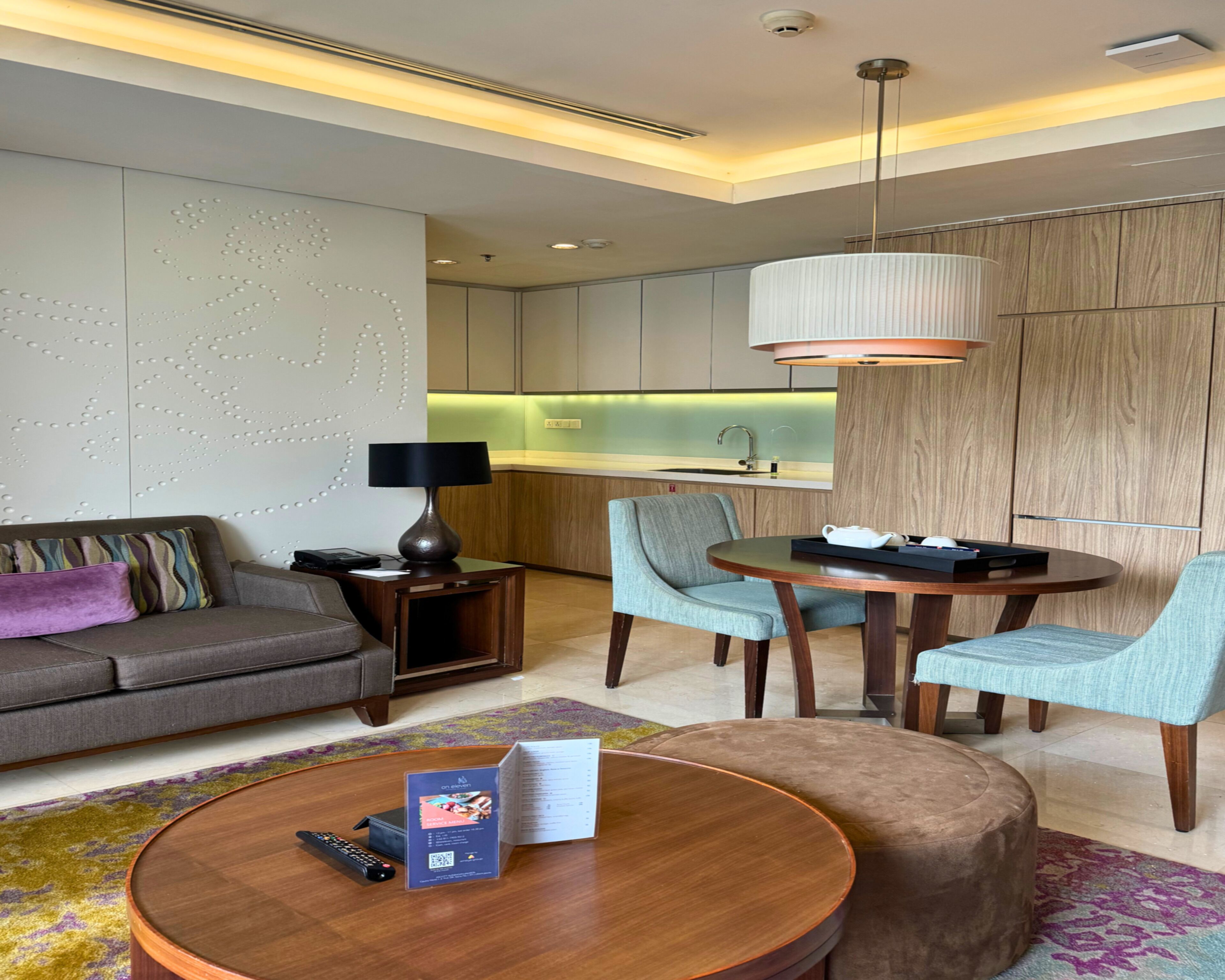 A modern hotel room with a sofa, a round coffee table, a dining table with chairs, a lamp, and a kitchen area in the background.