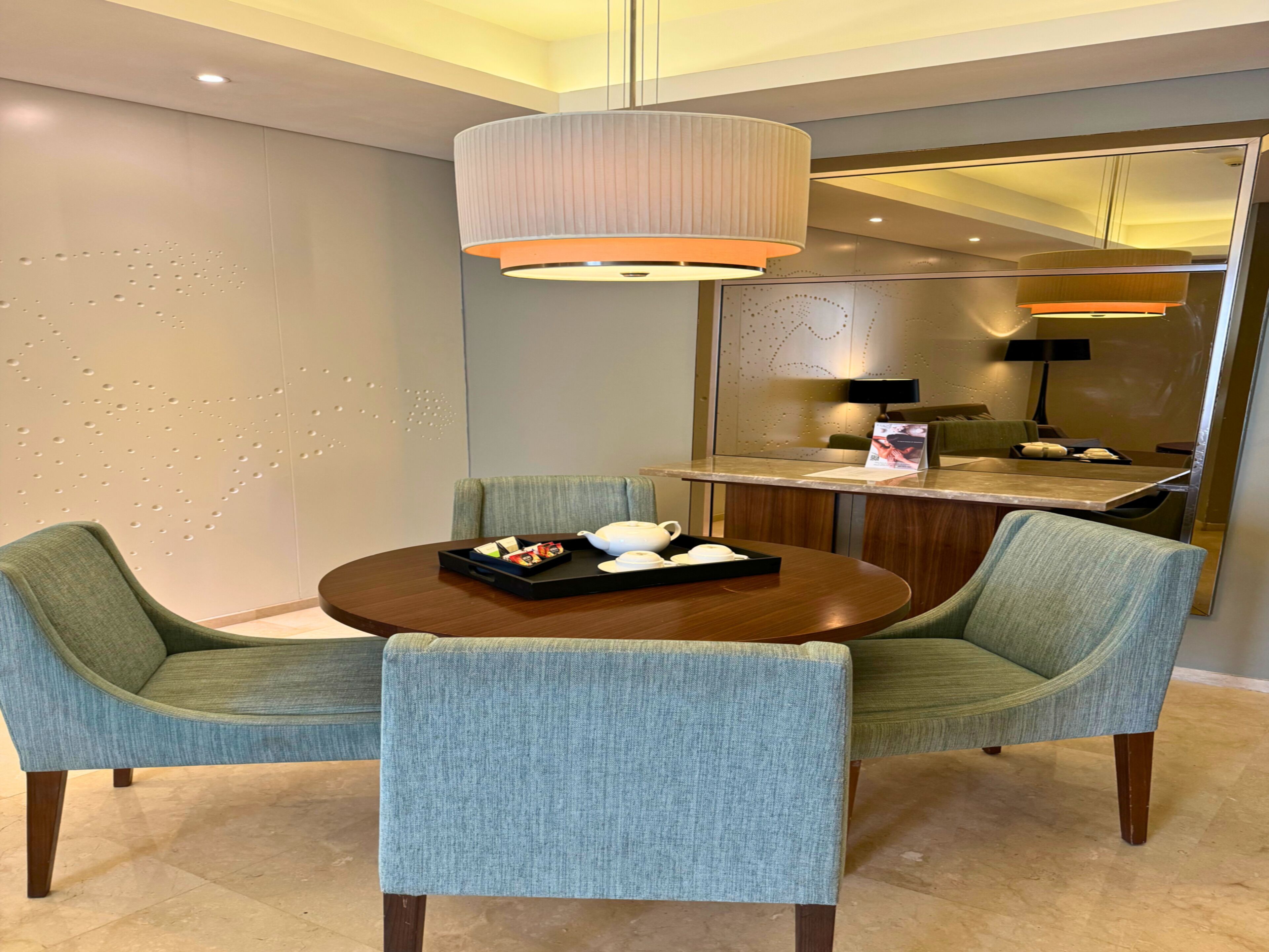 Un espace repas moderne avec une table ronde, deux chaises rembourrées, un mur décoratif orné d’un motif, un luminaire suspendu et un miroir qui reflète la pièce.