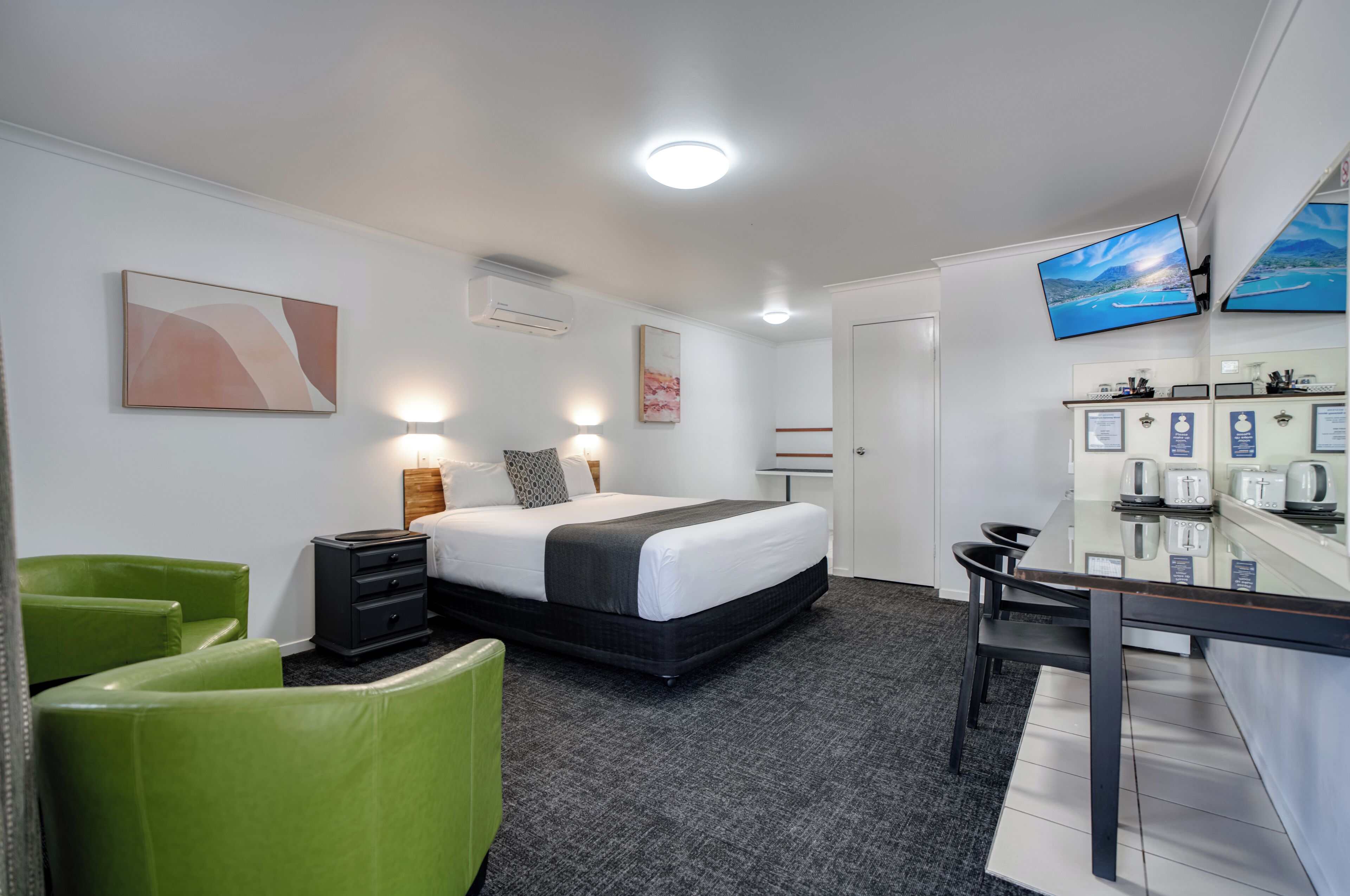 A hotel room with a bed, bedside tables, a desk, a chair, a flat-screen TV, and a wall-mounted air conditioning unit.