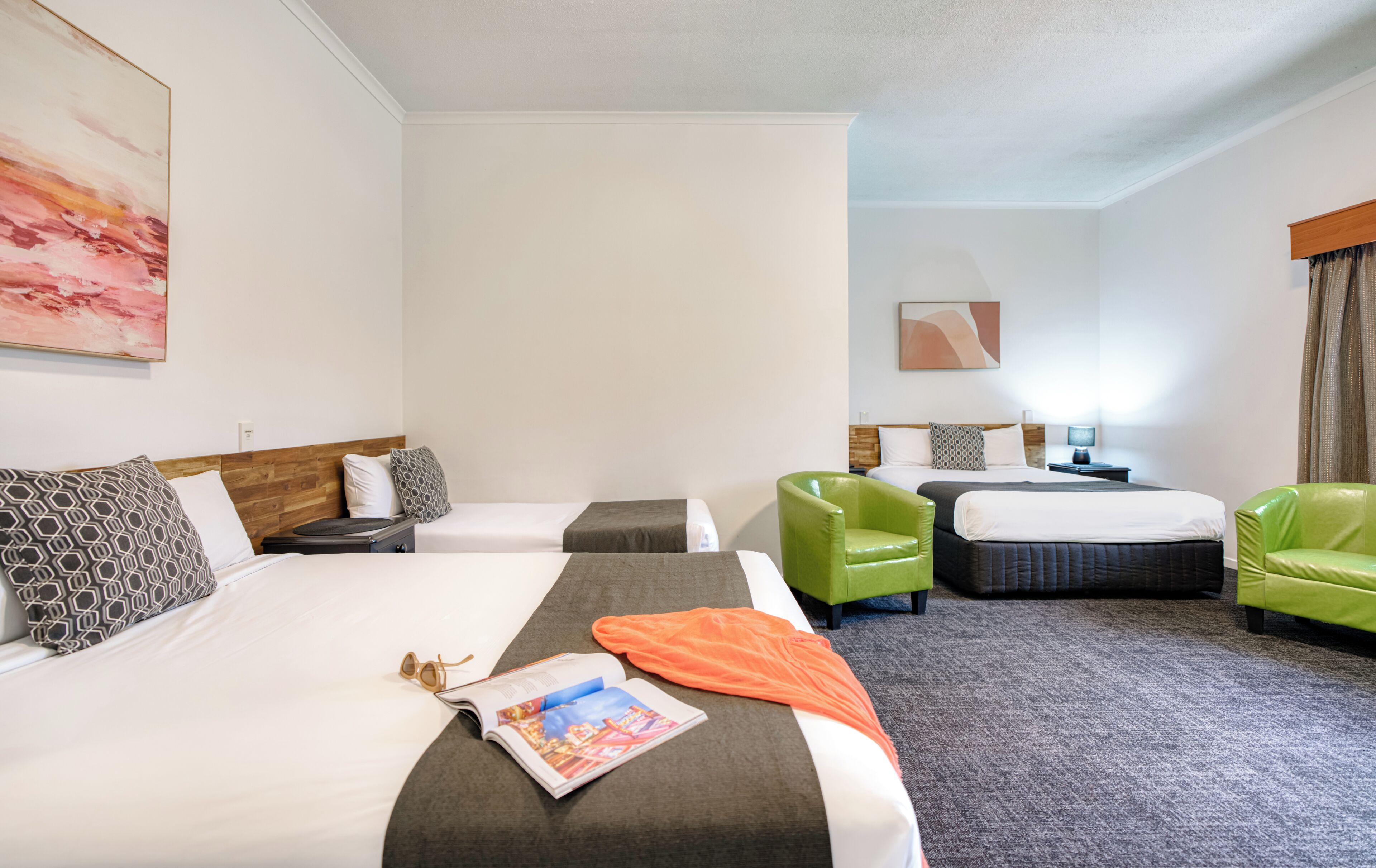 Habitación de hotel con dos camas, un sillón verde, una mesita de noche y un cuadro en la pared.