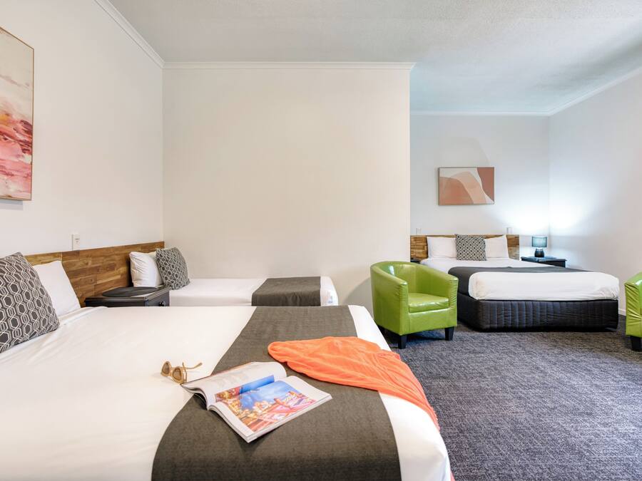 A hotel room with two beds, a green armchair, a nightstand, and a painting on the wall.