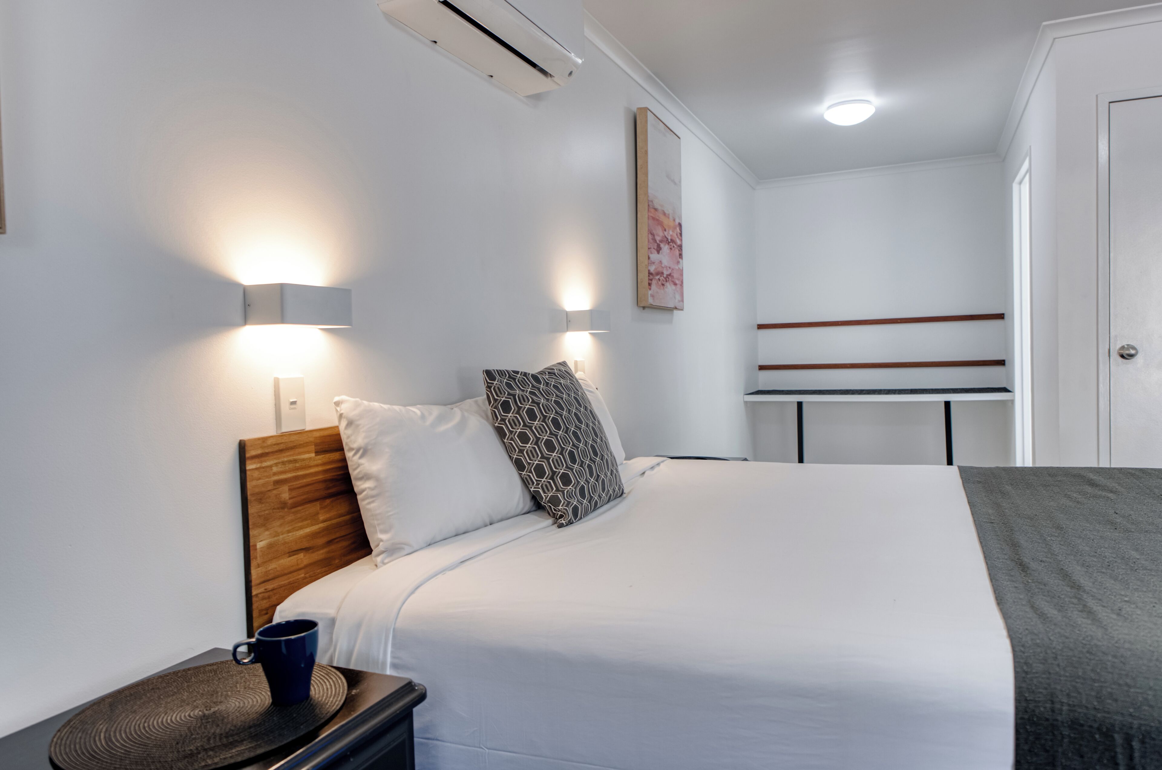 A hotel room with a bed, bedside table, lamp, and a bunk bed in the background.