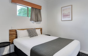 A hotel room with a bed, a window with curtains, a framed picture on the wall, and a small trash bin.