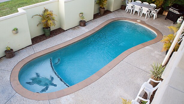 A rectangular swimming pool with a tiled edge, surrounded by a concrete deck. There are potted plants and a barbecue grill in the background. - Caboolture Gateway Motel (Caboolture)