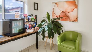 A room with a refrigerator stocked with snacks, a table with various food items, a green chair, and a large abstract painting on the wall. - Caboolture Gateway Motel (Caboolture)