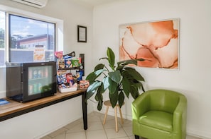 A room with a refrigerator stocked with snacks, a table with various food items, a green chair, and a large abstract painting on the wall. - Caboolture Gateway Motel (Caboolture)
