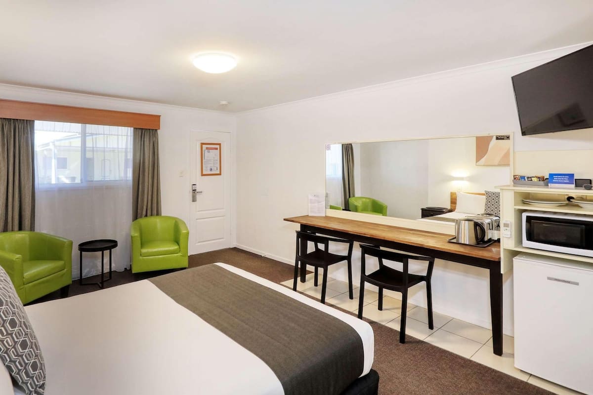 A hotel room with a bed, two green chairs, a desk with a mirror, a microwave, and a television.